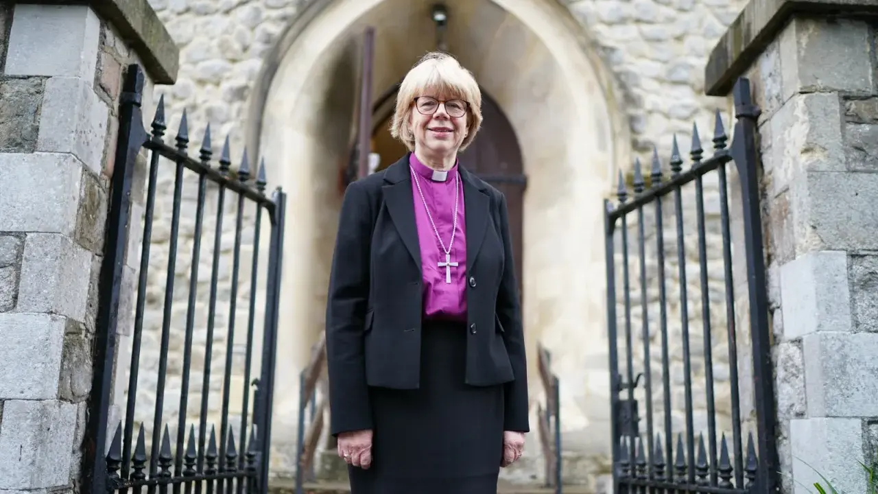 Eine Frau in geistlicher Kleidung, die an die Amtstracht des Erzbischofs von Canterbury erinnert, steht vor einer Kirche.