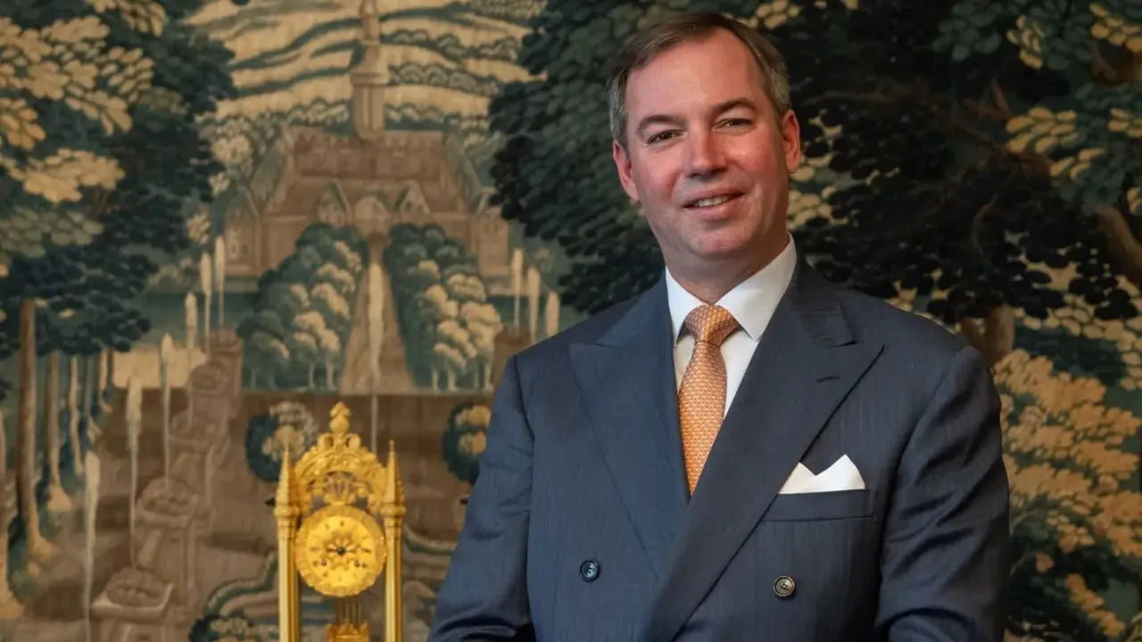 Ein Mann im Anzug, der wie ein Prinz von Frankreich aussieht, lächelt vor einem Wandteppich mit einer Landschaft und einer goldenen Uhr.