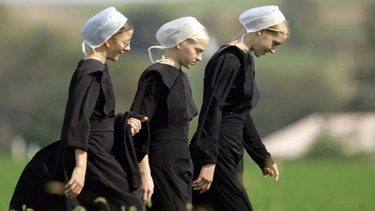 Drei junge Frauen in schwarzen Kleidern und weißen Hauben gehen über ein Feld. Sie verkörpern die Einfachheit, was bedeutet, amish zu leben.