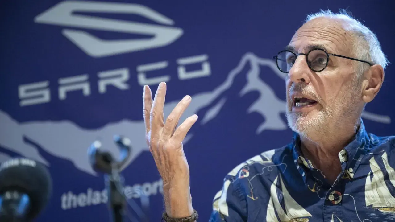 Philip Nitschke spricht mit erhobener Hand vor einem blauen Hintergrund mit dem SARCO-Logo.