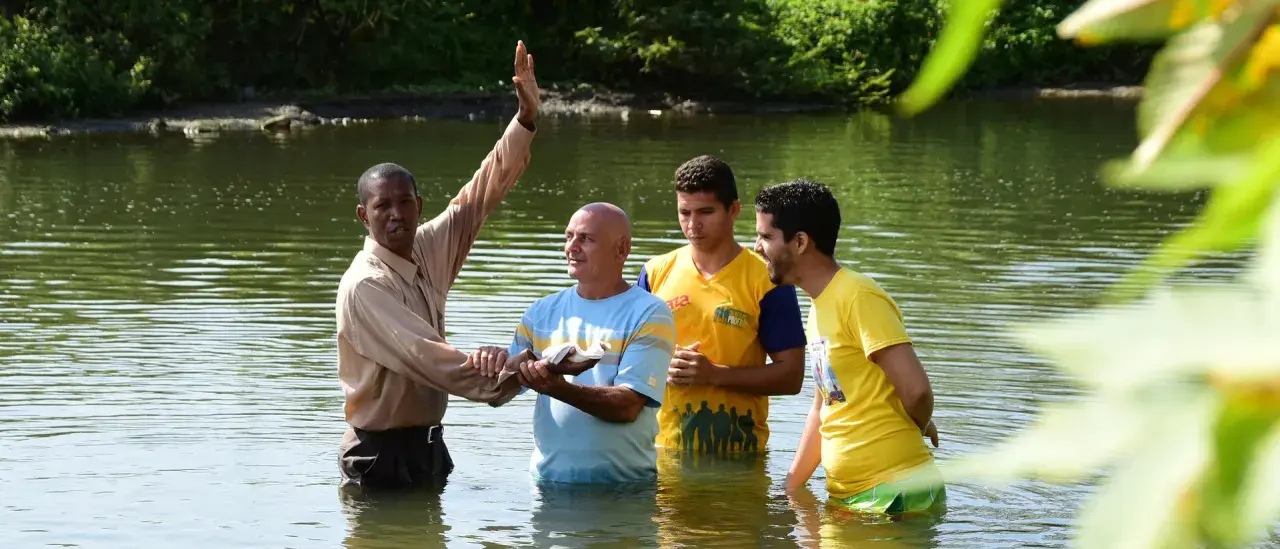 Ein Geistlicher führt eine Baptisten Taufe in einem Fluss durch. Drei Männer stehen im Wasser, während der Geistliche mit erhobener Hand segnet.