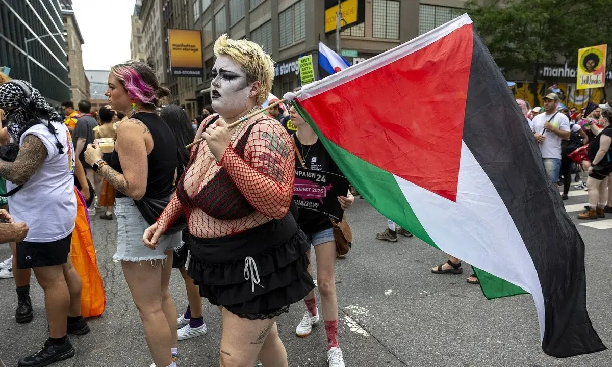 Queers for Palestine marschieren mit der palästinensischen Flagge. Eine Person mit auffälligem Make-up und Netzoberteil trägt die Flagge.