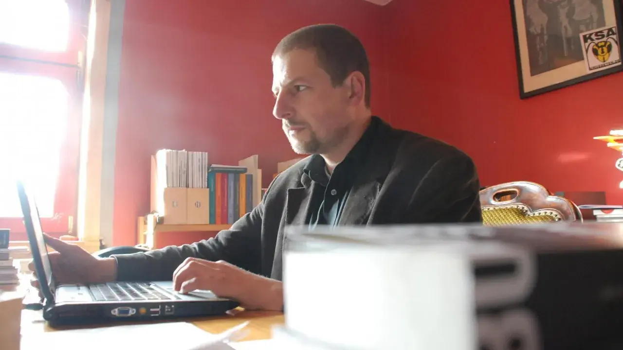 Mann arbeitet am Laptop im Büro des Antaios Verlags. Bücherregale im Hintergrund.