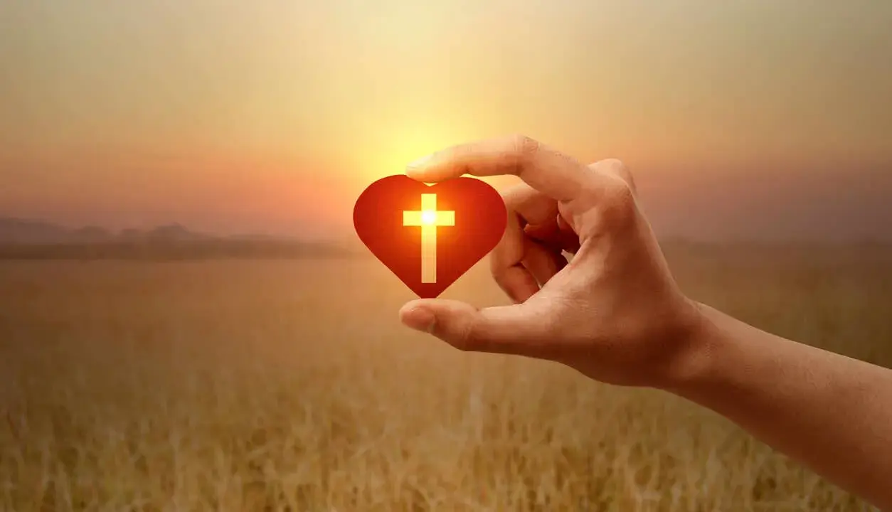 Hand hält Herz mit Kreuz, Symbol für christliche Werte im warmen Abendlicht über goldenem Feld.