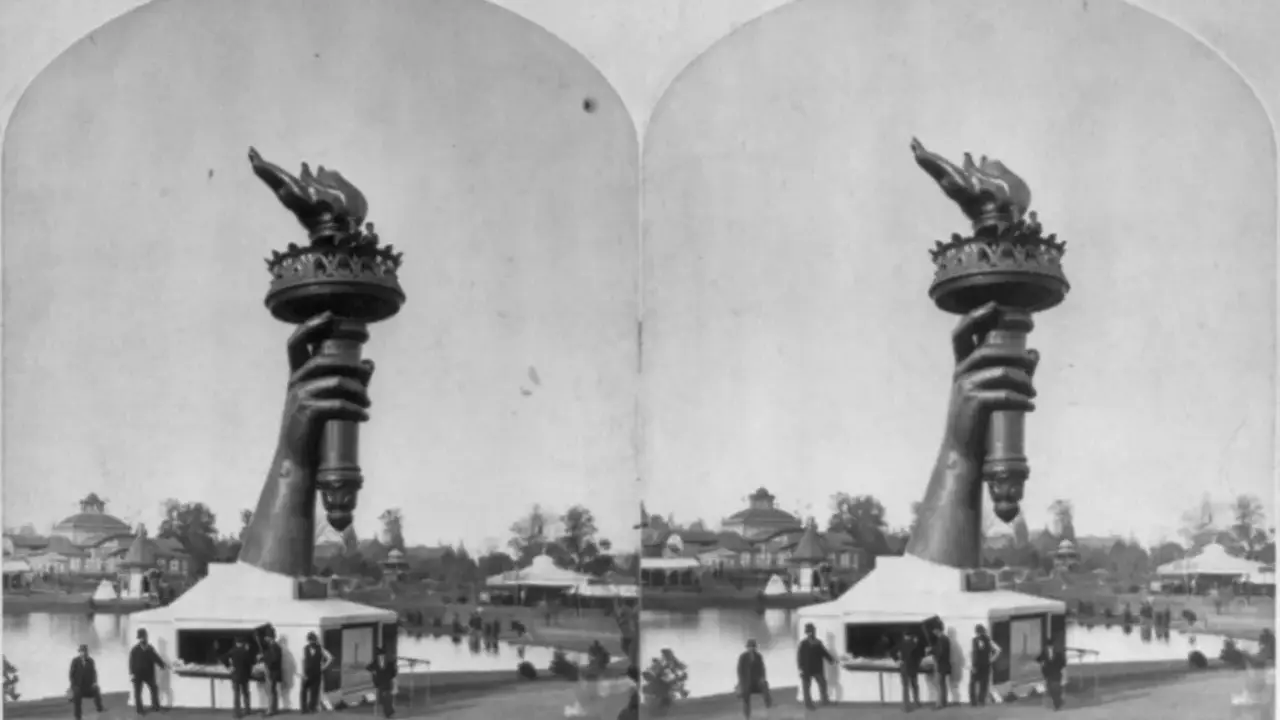Die Hand mit der Fackel der Freiheitsstatue, ein Werk der freiheitsstatue erbauer, ragt über einem Pavillon auf. Im Hintergrund ist eine Parklandschaft mit einem See und Gebäuden zu sehen.