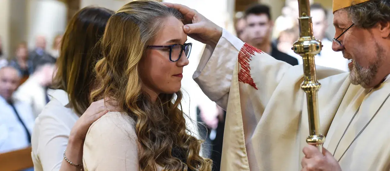 Ein Bischof legt einer jungen Frau die Hand auf die Stirn. Sie hat lange, blonde Haare und trägt eine Brille. Dies ist ein Moment, in dem sie ihren Glauben an Gott bekräftigt.