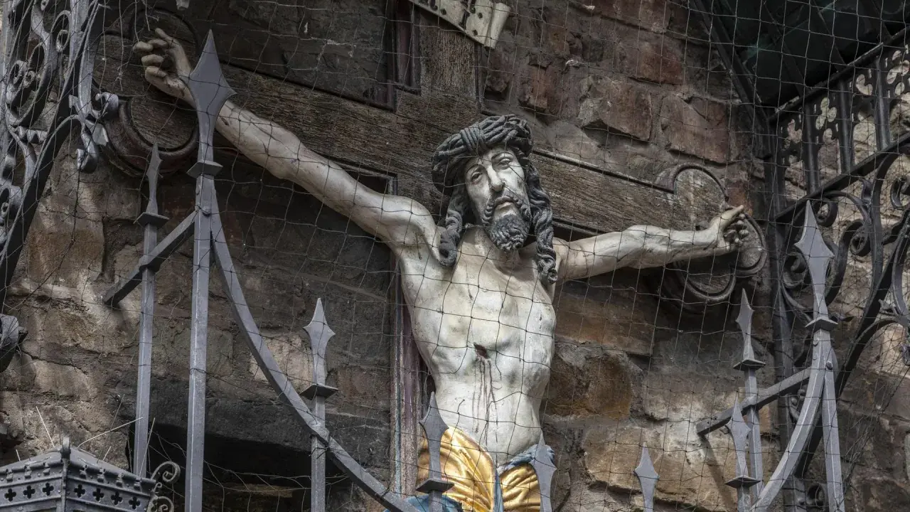 Skulptur Jesu am Kreuz, die an die Frage erinnert, warum wurde Jesus gekreuzigt. Die Darstellung ist durch ein Gitter geschützt.