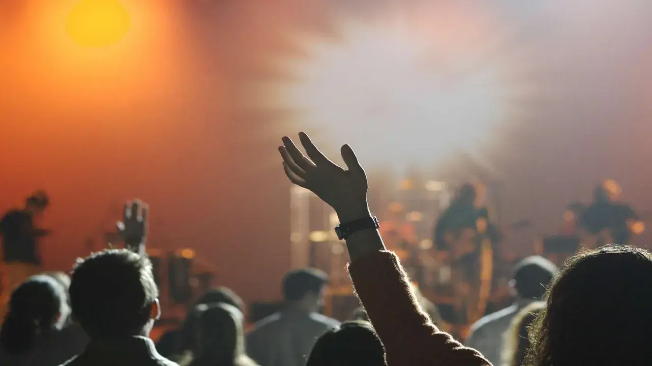 Eine Hand hebt sich im Scheinwerferlicht, während eine Menge bei einem Konzert jubelt. Dies spiegelt die lebendige Pfingstler Erziehung wider, die Gemeinschaft und Ausdruck fördert.