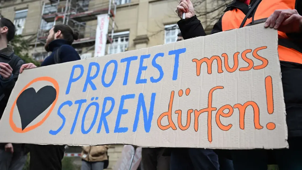 Menschen halten ein Schild mit der Aufschrift "PROTEST muss STÖREN dürfen!". Ein schwarzes Herz in einem orangen Kreis symbolisiert ziviler ungehorsam Beispiele.