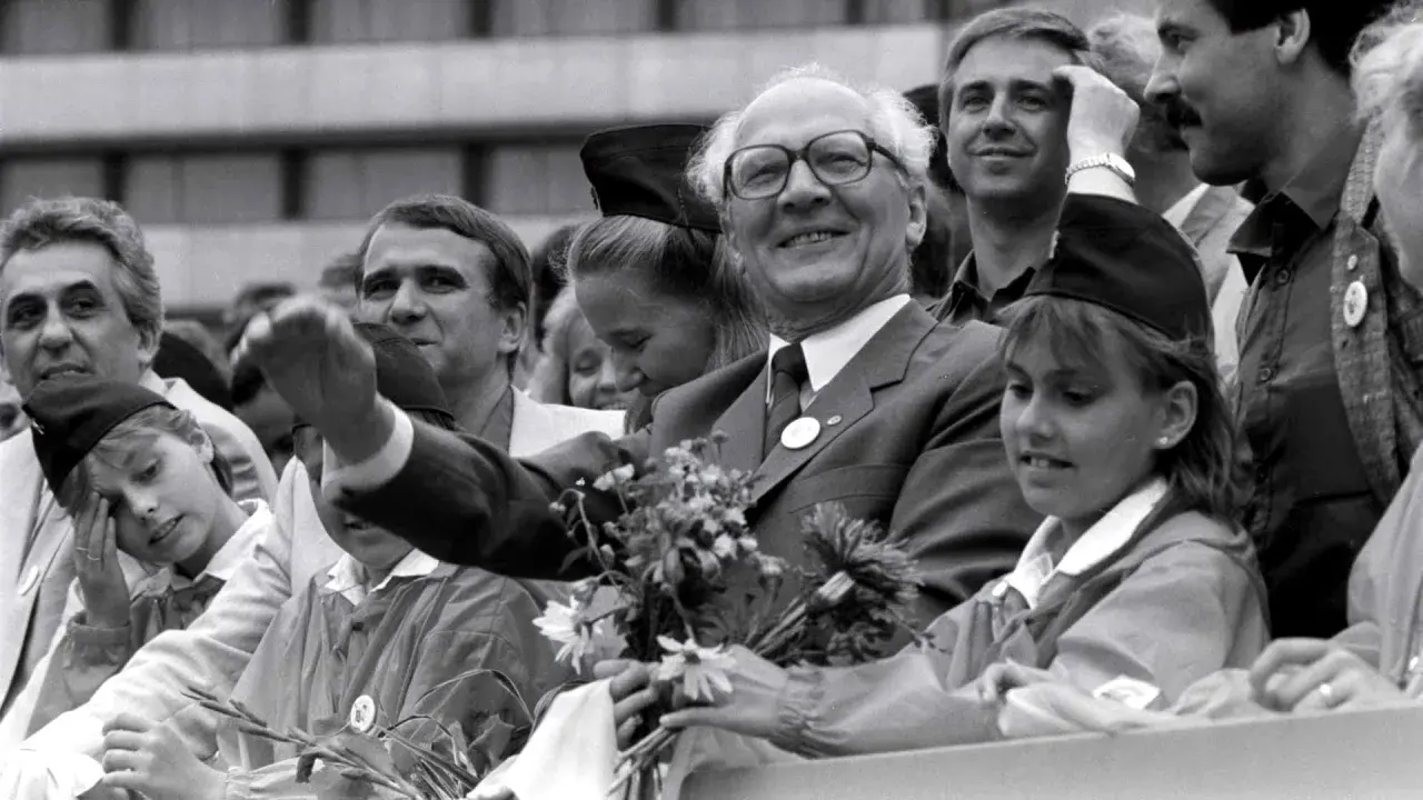 Ein Mann mit Brille winkt lächelnd, umgeben von Menschen. War die DDR eine Diktatur? Diese Szene zeigt eine öffentliche Veranstaltung, vielleicht ein Zeichen der Hoffnung.