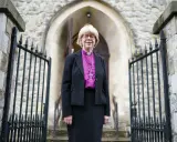 Eine Frau in geistlicher Kleidung, die an die Amtstracht des Erzbischofs von Canterbury erinnert, steht vor einer Kirche.
