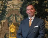 Ein Mann im Anzug, der wie ein Prinz von Frankreich aussieht, lächelt vor einem Wandteppich mit einer Landschaft und einer goldenen Uhr.