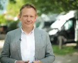 Johannes Sturm, ein Mann mittleren Alters mit freundlichem Lächeln, trägt ein Sakko und ein Hemd. Im Hintergrund sind Bäume und Fahrzeuge zu sehen.
