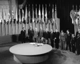 Männer in Anzügen stehen vor einer Reihe von Flaggen, während einer auf einem runden Tisch unterschreibt. Ein historisches Dokument, eine Charta, wird unterzeichnet.