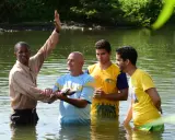 Ein Geistlicher führt eine Baptisten Taufe in einem Fluss durch. Drei Männer stehen im Wasser, während der Geistliche mit erhobener Hand segnet.