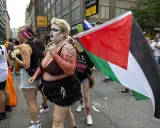 Queers for Palestine marschieren mit der palästinensischen Flagge. Eine Person mit auffälligem Make-up und Netzoberteil trägt die Flagge.