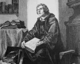 Martin Luther sitzt in seinem Studierzimmer und schreibt an seiner Bibelübersetzung. Bücher stapeln sich um ihn herum.
