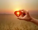 Hand hält Herz mit Kreuz, Symbol für christliche Werte im warmen Abendlicht über goldenem Feld.