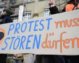 Menschen halten ein Schild mit der Aufschrift "PROTEST muss STÖREN dürfen!". Ein schwarzes Herz in einem orangen Kreis symbolisiert ziviler ungehorsam Beispiele.