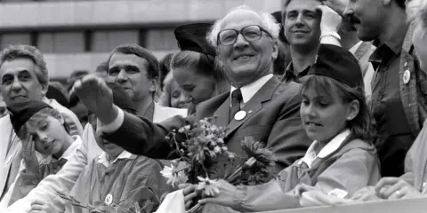 Ein Mann mit Brille winkt lächelnd, umgeben von Menschen. War die DDR eine Diktatur? Diese Szene zeigt eine öffentliche Veranstaltung, vielleicht ein Zeichen der Hoffnung.