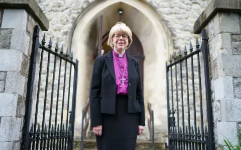 Eine Frau in geistlicher Kleidung, die an die Amtstracht des Erzbischofs von Canterbury erinnert, steht vor einer Kirche.