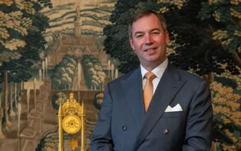 Ein Mann im Anzug, der wie ein Prinz von Frankreich aussieht, lächelt vor einem Wandteppich mit einer Landschaft und einer goldenen Uhr.