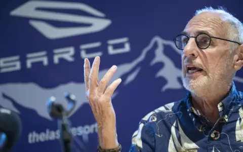 Philip Nitschke spricht mit erhobener Hand vor einem blauen Hintergrund mit dem SARCO-Logo.