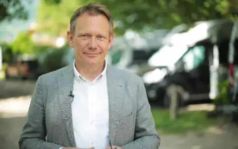 Johannes Sturm, ein Mann mittleren Alters mit freundlichem Lächeln, trägt ein Sakko und ein Hemd. Im Hintergrund sind Bäume und Fahrzeuge zu sehen.