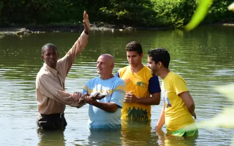 Ein Geistlicher führt eine Baptisten Taufe in einem Fluss durch. Drei Männer stehen im Wasser, während der Geistliche mit erhobener Hand segnet.