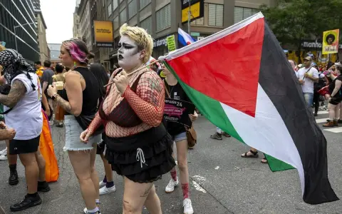 Queers for Palestine marschieren mit der palästinensischen Flagge. Eine Person mit auffälligem Make-up und Netzoberteil trägt die Flagge.