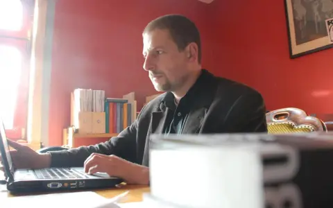 Mann arbeitet am Laptop im Büro des Antaios Verlags. Bücherregale im Hintergrund.
