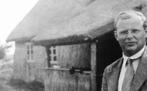 Ein Mann mit Brille steht vor einem Reetdachhaus. Er ist Dietrich Bonhoeffer, ein deutscher Theologe und Widerstandskämpfer.