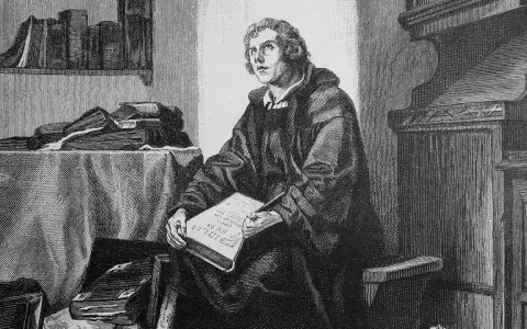 Martin Luther sitzt in seinem Studierzimmer und schreibt an seiner Bibelübersetzung. Bücher stapeln sich um ihn herum.