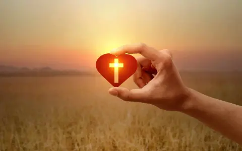 Hand hält Herz mit Kreuz, Symbol für christliche Werte im warmen Abendlicht über goldenem Feld.