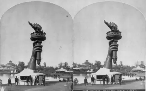 Die Hand mit der Fackel der Freiheitsstatue, ein Werk der freiheitsstatue erbauer, ragt über einem Pavillon auf. Im Hintergrund ist eine Parklandschaft mit einem See und Gebäuden zu sehen.