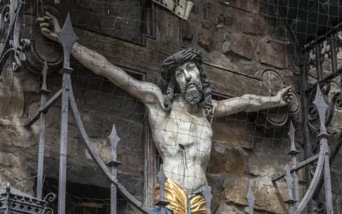 Skulptur Jesu am Kreuz, die an die Frage erinnert, warum wurde Jesus gekreuzigt. Die Darstellung ist durch ein Gitter geschützt.