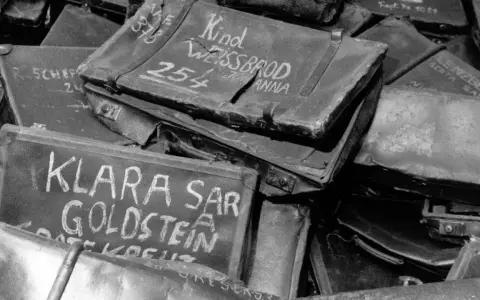 Stapel von alten Koffern, beschriftet mit Namen wie "Klara Goldstein" und "Kind Weissbrod Johanna". Diese Auschwitz Koffer sind ein trauriges Zeugnis der Vergangenheit.