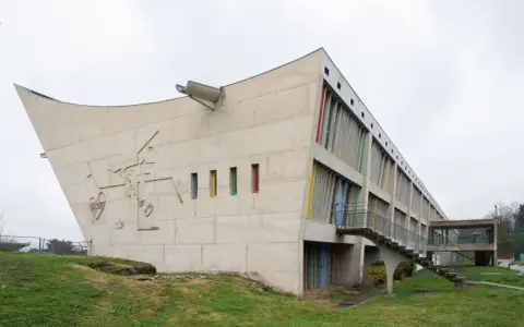 Betonbau mit bunten Fenstern und einer markanten, geschwungenen Form. Ein Meisterwerk des Architekten Le Corbusier.