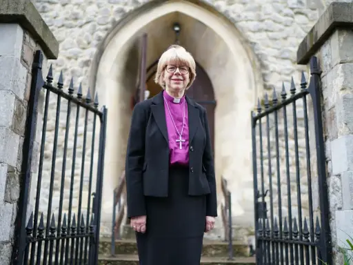Eine Frau in geistlicher Kleidung, die an die Amtstracht des Erzbischofs von Canterbury erinnert, steht vor einer Kirche.