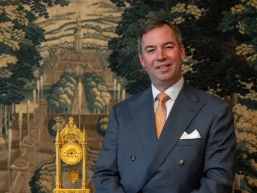 Ein Mann im Anzug, der wie ein Prinz von Frankreich aussieht, lächelt vor einem Wandteppich mit einer Landschaft und einer goldenen Uhr.
