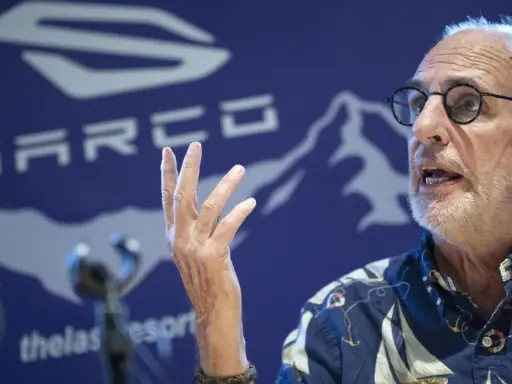 Philip Nitschke spricht mit erhobener Hand vor einem blauen Hintergrund mit dem SARCO-Logo.