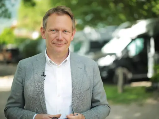 Johannes Sturm, ein Mann mittleren Alters mit freundlichem Lächeln, trägt ein Sakko und ein Hemd. Im Hintergrund sind Bäume und Fahrzeuge zu sehen.