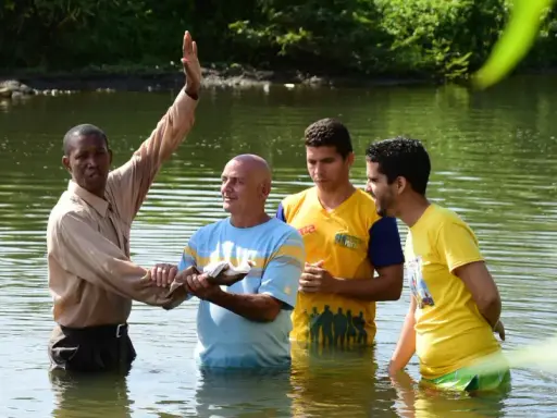 Ein Geistlicher führt eine Baptisten Taufe in einem Fluss durch. Drei Männer stehen im Wasser, während der Geistliche mit erhobener Hand segnet.