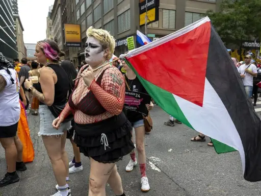 Queers for Palestine marschieren mit der palästinensischen Flagge. Eine Person mit auffälligem Make-up und Netzoberteil trägt die Flagge.