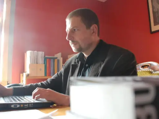 Mann arbeitet am Laptop im Büro des Antaios Verlags. Bücherregale im Hintergrund.