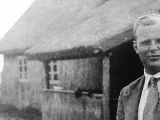 Ein Mann mit Brille steht vor einem Reetdachhaus. Er ist Dietrich Bonhoeffer, ein deutscher Theologe und Widerstandskämpfer.