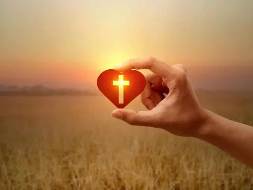 Hand hält Herz mit Kreuz, Symbol für christliche Werte im warmen Abendlicht über goldenem Feld.