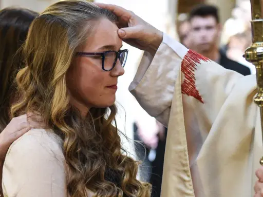 Ein Bischof legt einer jungen Frau die Hand auf die Stirn. Sie hat lange, blonde Haare und trägt eine Brille. Dies ist ein Moment, in dem sie ihren Glauben an Gott bekräftigt.