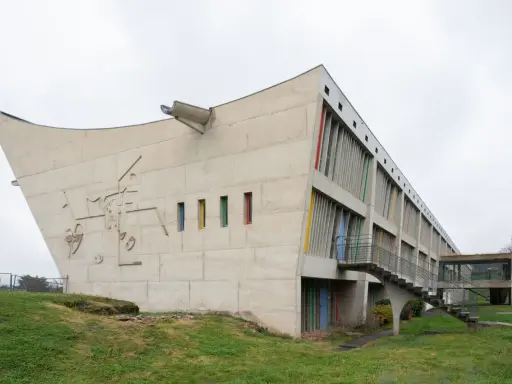 Betonbau mit bunten Fenstern und einer markanten, geschwungenen Form. Ein Meisterwerk des Architekten Le Corbusier.