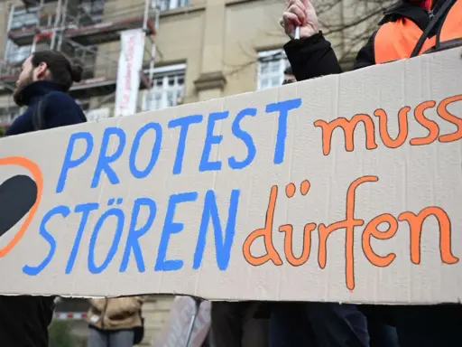 Menschen halten ein Schild mit der Aufschrift "PROTEST muss STÖREN dürfen!". Ein schwarzes Herz in einem orangen Kreis symbolisiert ziviler ungehorsam Beispiele.